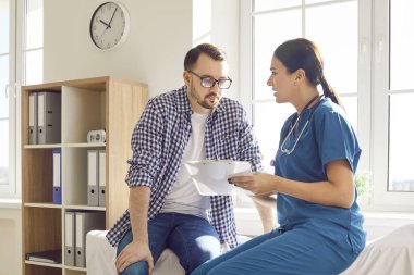 Kadın doktor ve erkek hasta hastane ofisinde sohbet ediyorlar. Gülüşleri sadece tıbbi uzmanlığı değil aynı zamanda kaliteli hasta bakımını tanımlayan insan dokunuşunu da yansıtıyor..