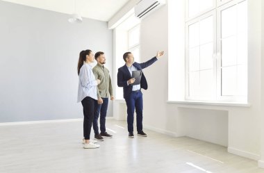 Olgun bir emlakçı, yeni ev ya da dairede potansiyel alıcılarla duran bir emlakçı. Onlara pencereden harika bir manzara gösteriyor. Genç bir çift boş odada duruyor, yeni bir ev alıyorlar..