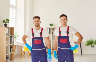 Profesyonel temizlik servisinden gülümseyen iki hademenin mutlu portresi ellerinde deterjan tutarken kameraya neşeli görünüyor. Ev işleri, ev işleri ve ev idaresi kavramı.