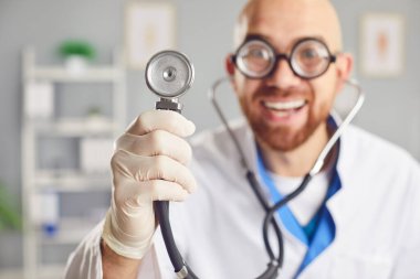 Klinikte steteskopla dikilen beyaz üniformalı komik, genç, mutlu bir erkek doktorun portresi. Gülümseyen doktor kameraya neşeli bakıyor. Seçici odak.