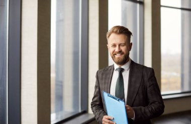 Kendine güvenen bir iş adamı ya da resmi takım elbiseli bir emlakçı boş dairelerde elinde belgelerle kameraya neşeli bir şekilde bakıyor. İpotek ve iş portresi konsepti taşınıyor