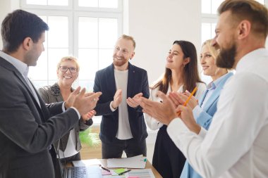 Meslektaşlar başarılı bir projeyi kutlamak için parlak bir ofiste toplanırlar. Sunucuyu alkışlıyor ve gülümsüyorlar, takım çalışması ve pozitif iş yeri kültürü sergiliyorlar..