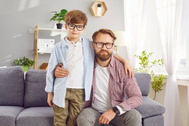 Kendine güveni tam genç bir babanın portresi. Kızıl saçlı, gözlüklü küçük çocuğu içeride sarılıyor. Baba oğul evdeki kanepede oturan kameraya bakıyor. Babalar günü ve babalık kavramı..