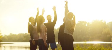 Spor kıyafetli bir grup insan göl kıyısında bir çember oluşturarak parktaki gün batımında doğa çalışmasından sonra ellerini kaldırdılar. Açık hava eğitimi, spor konsepti. Pankart.