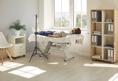 Genç sakallı adam, kanında serum infüzyonu ve vitamin tedavisi görürken pencerenin yanındaki klinikte kanepede yatıyor. Enjeksiyon tedavisi gören kişi. Sağlık hizmetleri kavramı.