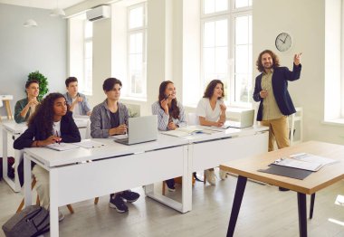 Lisedeki erkek arkadaş canlısı öğretmen. Öğrenciler ve sınıf arkadaşları sıralarında oturmuş hocalarını dinliyor ve modern sınıfta dizüstü bilgisayarla çalışıyorlar. Eğitim kavramı.