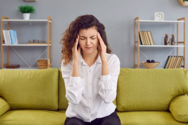 Evdeki kanepede oturan kıvırcık saçlı, baş ağrısı çeken genç bir kadının portresi. Hasta kız stres, migren ya da depresyon hissediyor. Sağlık ve Tıp Konsepti.