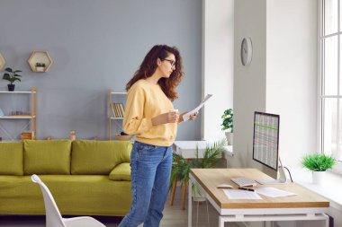 Genç, kıvırcık saçlı iş kadını, gözlüklü muhasebeci, iş yerinin masasının yanında elinde masalarla ya da elinde kahve ya da çayla duruyor..