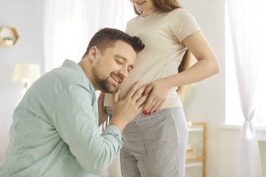 Mutlu koca, hamile kadının karnındaki bebeği evde, oturma odasında dinliyor. Genç bir çift ve bebek bekleyen müstakbel ebeveynler. Ebeveynlik, annelik ve hamilelik kavramı.