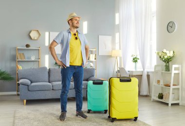 Mutlu gülümseyen adamın uzun bir portresi. Yaz tatiline hazır bavullarıyla oturma odasında duruyor. Neşeli erkek turist tatile çıkıyor. Seyahat ve seyahat kavramı.