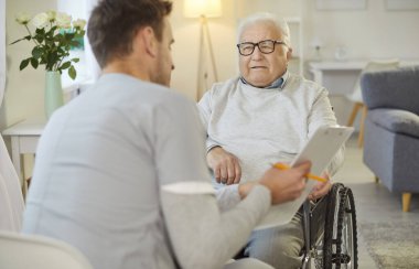 Genç, dost canlısı erkek hemşire ya da bakıcı yaşlı, tekerlekli sandalyedeki engelli hastaya test sonuçları gösteriyor ve tıbbi klinikte ona reçete yazıyor. Sağlık ve hemşirelik kavramı.