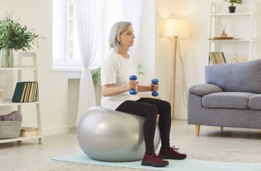 Yaşlı bir kadın spor egzersizleri yapıyor. Elinde dambıllarla topun üzerinde oturuyor. Olgun bir kadın evdeki oturma odasında egzersiz yapıyor. Sağlıklı yaşam tarzı, fitness ve spor kavramı.