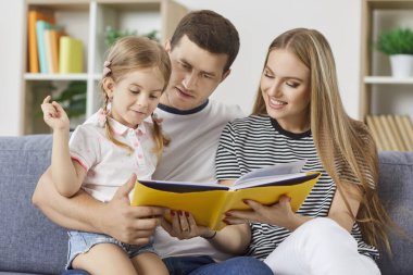 Evdeki kanepede oturan küçük tatlı kızlarıyla kitap okuyan mutlu bir aile. Gülümseyen ebeveynler sevimli çocuklarıyla vakit geçirip gülümsüyorlar. Birlikte boş zaman konsepti.