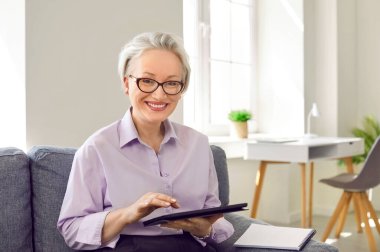 Orta yaşlı mutlu bir iş kadınının portresi. Mutlu, kendine güvenen, kendine güvenen, güzel, beyaz saçlı, şık gözlüklü bir kadın. Ofisteki koltukta oturuyor, tablet bilgisayar kullanıyor ve gülümsüyor.