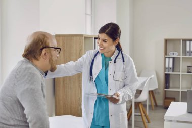 Beyaz önlüklü güzel, gülümseyen kadın doktor, tıbbi kurum ofisinde olumlu haberler veren gözlüklü yaşlı adamla saygılı bir şekilde iletişim kuruyor. Psikolojik sağlık konsepti.