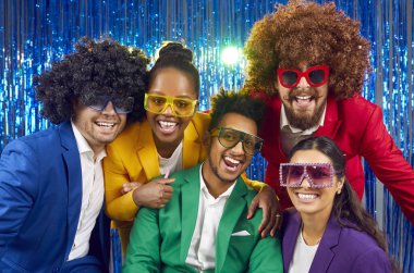 Neşeli, neşeli insanlar farklı yeşil, kırmızı, mor kostümler, parti gözlükleri ve komik peruklar takıyor gülümsüyor ve parlak mavi folyo fotofon arka planında kameraya bakıyor.