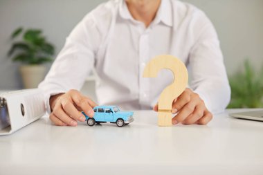 Ofis masasında oturan bir araba sigortacısı elinde küçük mavi bir oyuncak otomobil mankeni ve ellerinde ahşap bir soru işareti tutarken görüntülenmiş. Araba sigortası konsepti