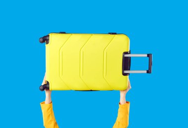 Elinde bavulunu taşıyan genç bir kadın mavi arka planda izole edilmiş, kırpılmış bir stüdyo fotoğrafı. Modern sarı tekerlekli bavuluyla uçakla ya da trenle seyahat eden mutlu bir turist.