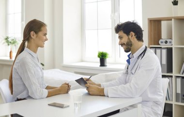 Doktorun, muayenehanesinde masada oturan bir hastayla güzel bir sohbet edişinin yan görüntüsü. Modern klinik ya da hastane ziyareti sırasında doktorları dinleyen mutlu genç kadın.