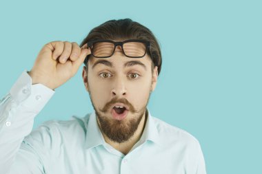 Afallamış genç adamın stüdyo fotoğrafı. Beyaz ofis gömleği içinde sakallı ve bıyıklı yakışıklı bir adam ağzını açar, gözlüklerini kaldırır ve komik, şaşırmış yüz ifadesi olan bir şeye bakar.