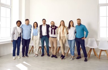 Kameraya bakarken gülümseyen ve sarılan neşeli insanların portresi. Gündelik ve şık giyimli neşeli çok ırklı meslektaşlar parlak ışık ofisinde ya da lobide sıraya giriyorlar..
