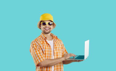 Mutlu adam dizüstü bilgisayarını yaz turu, otel rezervasyonu ya da tatildeyken internette çalışmak için kullanıyor. Yaz panamalı, gömlekli, güneş gözlüklü ve elinde dizüstü bilgisayarla açık mavi arka planda neşeli bir adam..