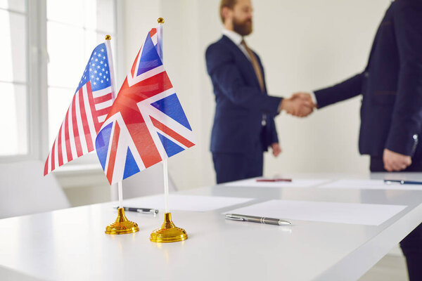 Political cooperation. Small flags of Britain and USA against background of handshake of politicians or businessmen. Close up of Us and UK flags on background of men in suits who approved agreement.