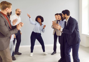 Neşeli iş arkadaşları alkışlarken Afrikalı Amerikalı iş kadını dans ediyor. Bir grup iş adamı iş yerlerinde takım oluşturma çalışmalarına katılıyor. Kavram oluşturma etkinliği, zafer dansı