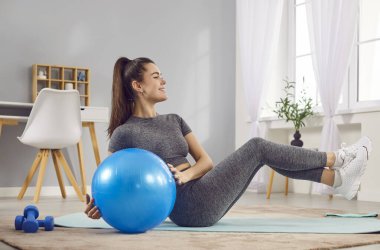 Spor formundaki genç bir kadın sabah sporu yapıyor. Oturma odasında topla tek başına egzersiz yapıyor. Spor kıyafeti giyen bir kız.