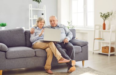 Yaşlı, güler yüzlü bir çift evdeki kanepede aileleriyle video görüşmesi yapıyor. Yaşlı bir adam ve kadın modern bir dizüstü bilgisayar kullanarak internette konuşuyorlar. Emeklilik ve iletişim teknolojisi kavramı.