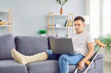 Evde dizüstü bilgisayarda çalışan engelli genç adam koltuk değnekleriyle oturuyor. Kırık bacağı olan yaralı bir erkek içeride rehabilitasyonda bilgisayar kullanıyor. Rehabilitasyon kavramı.