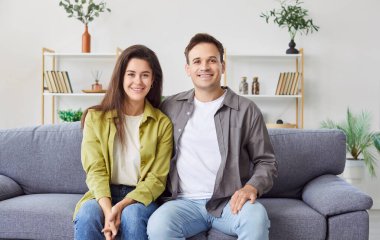 Mutlu genç karı ve kocanın mutlu aile portresi kanepede oturuyor, gülümseyen çift sevgi ve şefkat göstermek için sarılıyor, hafta sonları evde birlikte rahatlıyor, yeni bir daireye taşınmanın keyfini çıkarıyor, kameraya bakıyor.