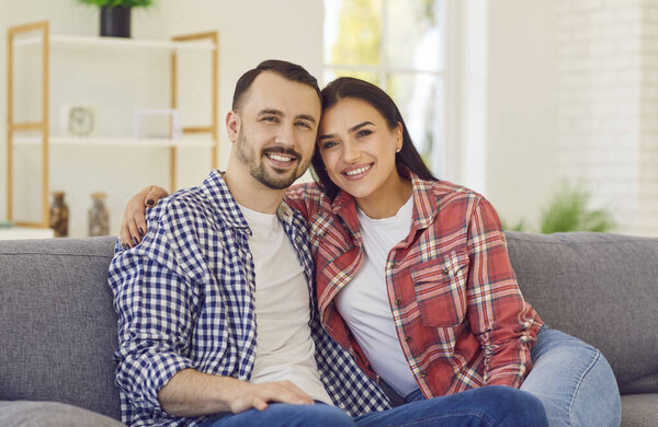 Портрет молодой счастливой супружеской пары, сидящей дома на диване. Улыбающиеся семьянин и женщина на диване обнимаются и смотрят в камеру. Концепция гармонических отношений.