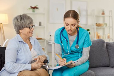 Steteskoplu genç hemşire tekerlekli sandalyedeki gülümseyen yaşlı kadınla konuşurken not alıyor. Konsültasyon ya da sağlık uzmanı ev ortamındaki şikayetleri dinliyor.