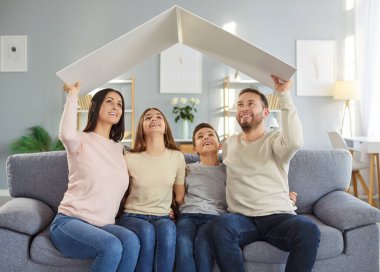Mutlu, gülümseyen bir ailenin portresi. İki çocuklu bir çocuk ve iki kız evdeki kanepede oturmuş kucaklaşıyorlar ve yukardaki evi tutuyorlar. Sigorta, ipotek, güvenlik ve koruma kavramı.