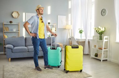 Sahil şapkalı mutlu genç adam yaz tatiline hazır bavullarıyla oturma odasında duruyor. Erkek turist tatile çıkıyor. Seyahat ve seyahat kavramı.