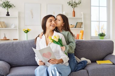 Ergen kız, ona güzel hediyeler vererek anneler gününü kutluyor. Çocuk annesine arkadan sarılıyor, onu öpüyor ve kalp ve bir buket sarı lale ile süslenmiş el yapımı bir kart veriyor..