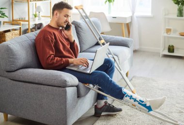 Kırık bacaklı genç adam dizüstü bilgisayarda çalışıyor cep telefonuyla konuşuyor koltuk değnekleriyle oturuyor. Erkek yaralı içeride rehabilitasyonda bilgisayar kullanıyor..