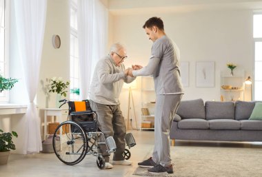 Yaşlılar yurdunda tekerlekli sandalyeden kalkması için yaşlı bir hastaya yardım eden genç bir erkek hemşirenin portresi. Fizyoterapist ya da sağlık görevlisi rehabilitasyondaki engelli yaşlı adama yardım ediyor..
