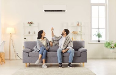 Evdeki kanepede battaniyeye sarılı oturan ve uzaktan kumandayla modern klimada rahat bir sıcaklık ayarlayan neşeli bir çift..