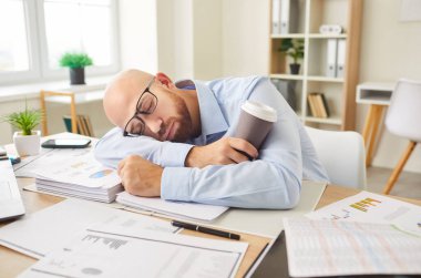 Ofisteki belgelerle kaplı bir masada uyuyan adam işten yorgun düşmüş. Kahve fincanı yorucu bir çalışma ortamında yorgunluğun etkisini vurguluyor..