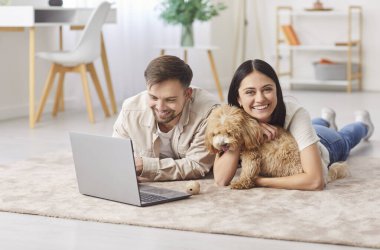 Çevrimiçi dizüstü bilgisayar izleyen genç bir çift, evde sevimli bir evcil hayvan yavrusu olan mutlu bir aile, eşler halı üzerinde küçük soylu köpekle eğleniyor, modern oturma odasında boş zaman aktivitesi yapıyor.