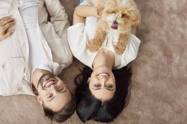 Evcil köpeği olan çocuksuz ve çocuksuz bir aile, sıcak bir ilişki uyumu içinde. Yukarıdan gülümseyen genç adam ve kadının bej renkli tüylü arkadaşıyla kahverengi halıda uzandığı kapalı bir çekim.