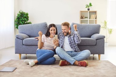 Genç aile, internette para kazandıklarında şanslarına inanamıyor. Kadın ve erkek mobil uygulamadan para kazanıyor. İki mutlu, heyecanlı insan koltuğun yanında oturuyor, cep telefonuna bakıyor ve bağırıyor.