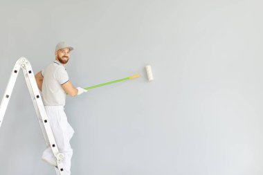 Gülümseyen bir ressamın portresi yenileme sırasında bir duvarda paten fırçasıyla çalışır. Profesyonellik, işteki neşe ve ev içini geliştiren inşaat süreci..