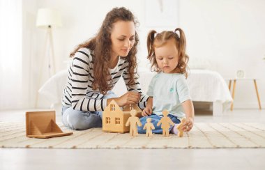Küçük kız annesiyle halıda oturuyor ve çatısı olmayan ahşap evle oynuyor ve güneşli yatak odasında tahta insan figürleriyle oynuyor. Aile mülkü ve gayrimenkul satın alma kavramı.