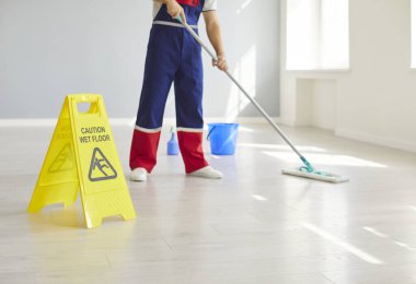 Düşük açılı, erkek hademenin bacaklarını paspas ile temizlerken çekilmiş bir fotoğrafı. Üniformalı adam temizlik işinde çalışıyor. Boş odayı ya da ofisi paspaslıyor..