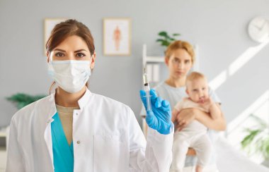 Tıbbi klinikte durup anne ve küçük bebeğin elinde şırıngayla kameraya bakan genç bir kadın doktor ya da hemşire portresi. Çocuk Aşılama Konsepti.