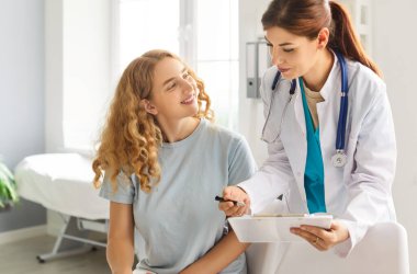 Mutlu gülümseyen genç kadın hasta, klinikteki muayene sırasında ofisinde oturan randevusuyla birlikte elinde rapor tutan dost canlısı kadın doktor terapistiyle konuşuyor. İlaç ve sağlık hizmetleri kavramı