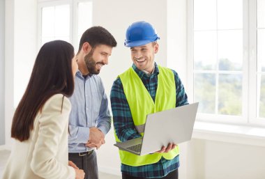 Mavi şapka ve güvenlik yeleği giyen neşeli inşaat işçisi yeni evlerindeki genç çifte dizüstü bilgisayardaki detayları sunuyor. Mutlu adam ve kadın inşaatın ilerlemesini müteahhitlerle tartışıyor..